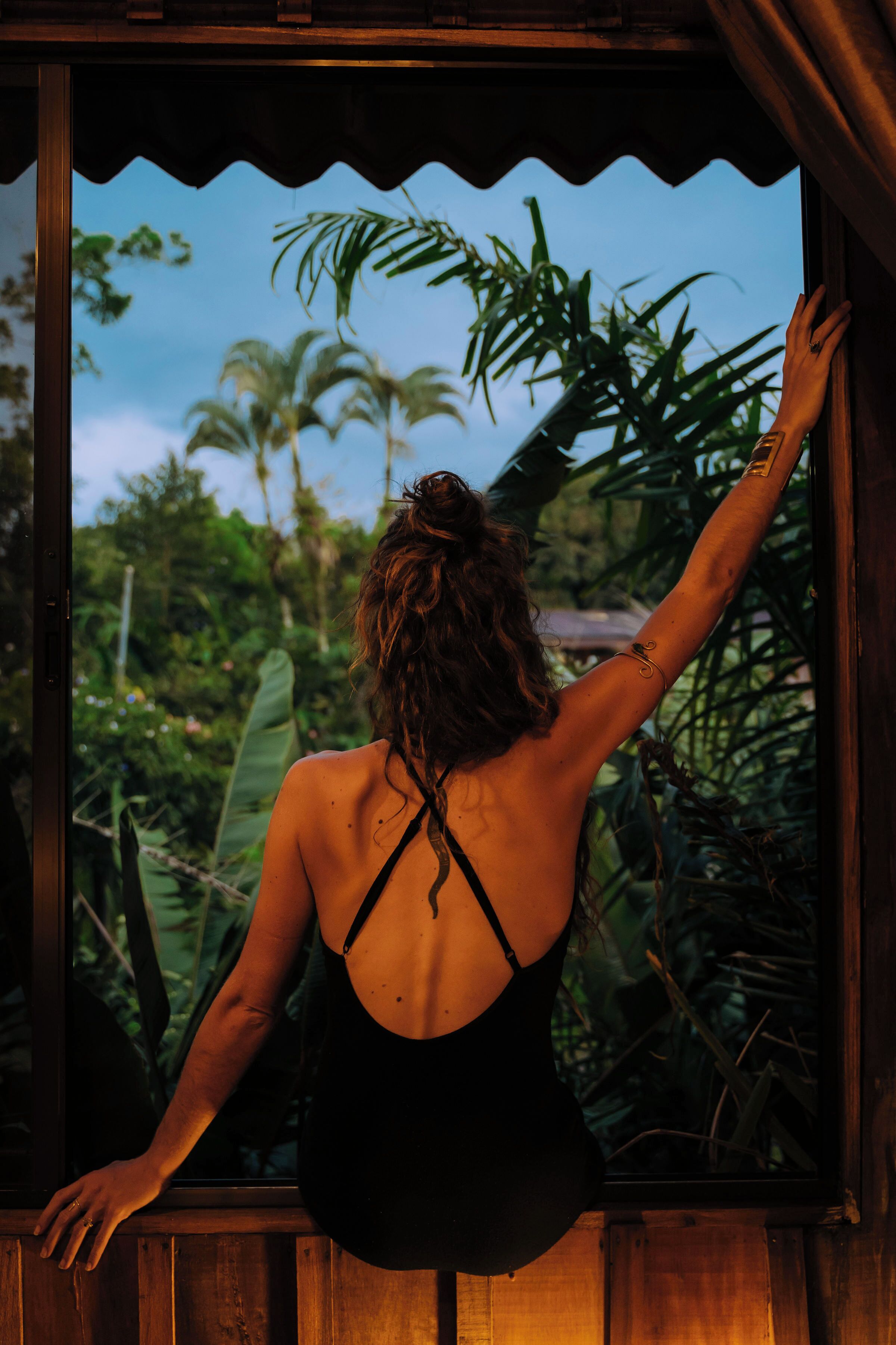 Une personne vêtue d’un maillot de bain noir, appuyée sur un cadre de fenêtre en bois, avec une vue sur la végétation tropicale.