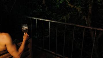 A person holding a glass of beer on a wooden deck with a metal railing, surrounded by dense foliage.