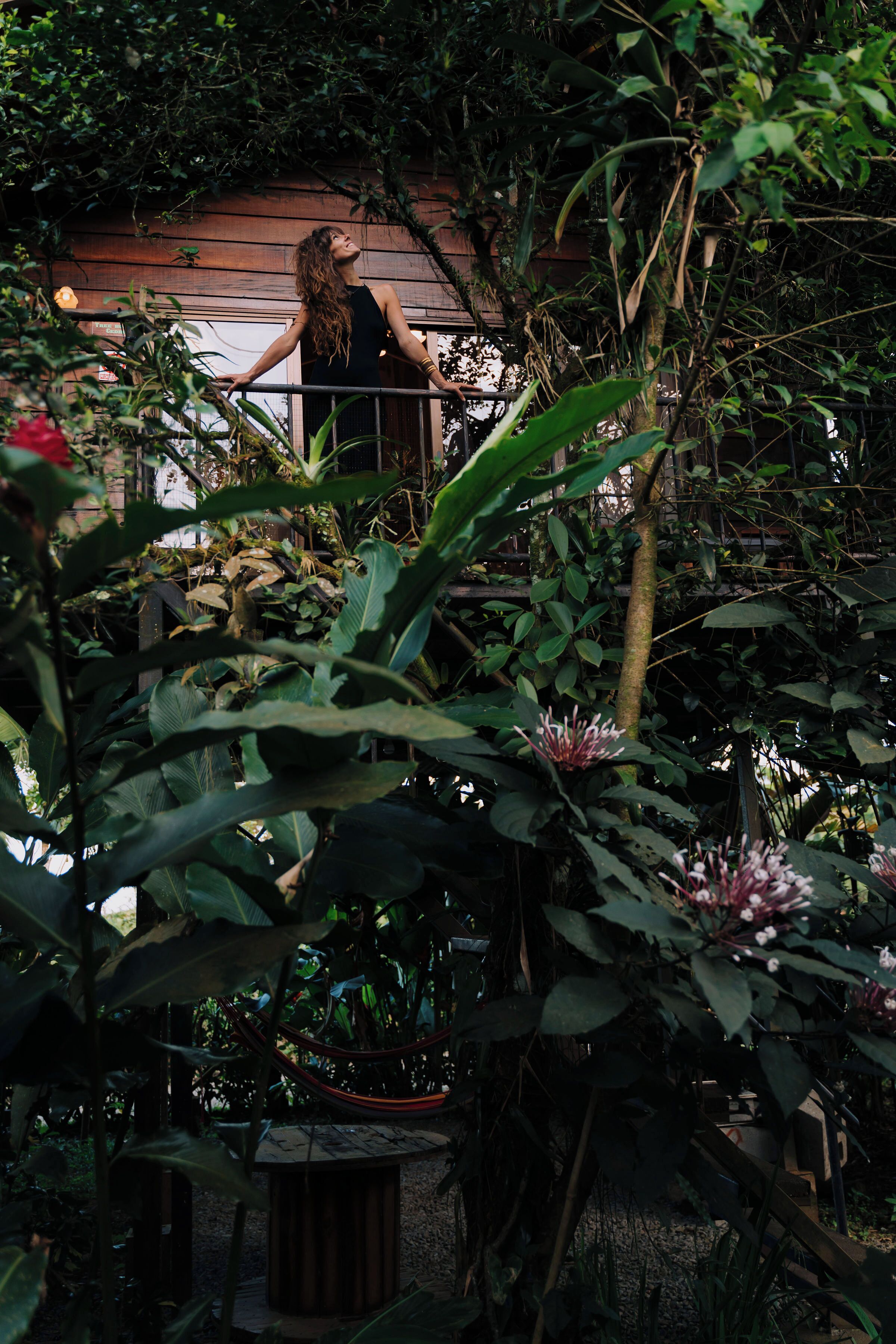 A woman standing on a balcony with lush greenery and a wooden structure in the background.