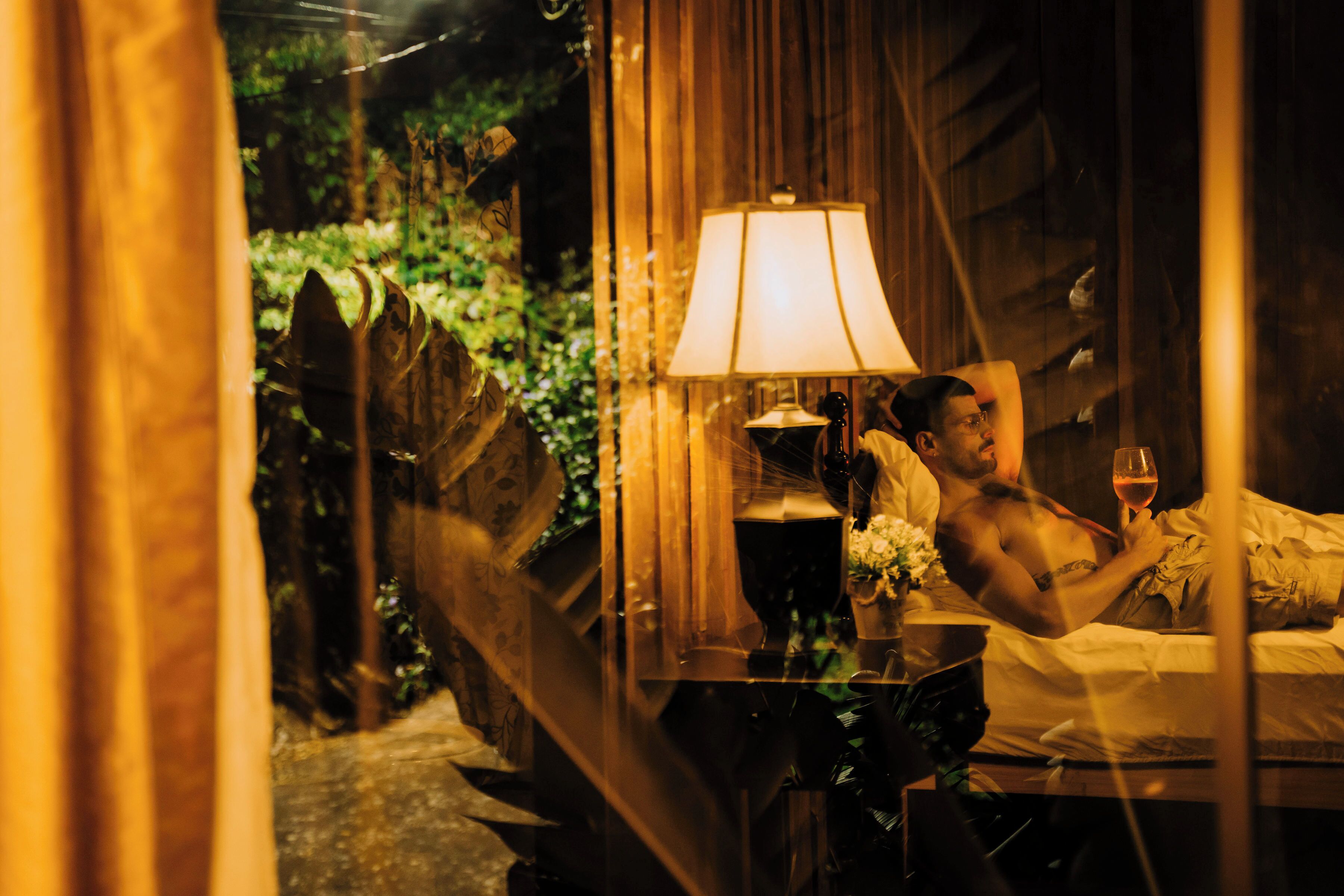 A dimly lit room with a bed, a bedside table with a lamp, and a window overlooking greenery.
