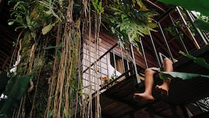 A person sitting on a wooden bench with a view of a wooden structure and hanging plants. - Las Cabanas (Venecia)