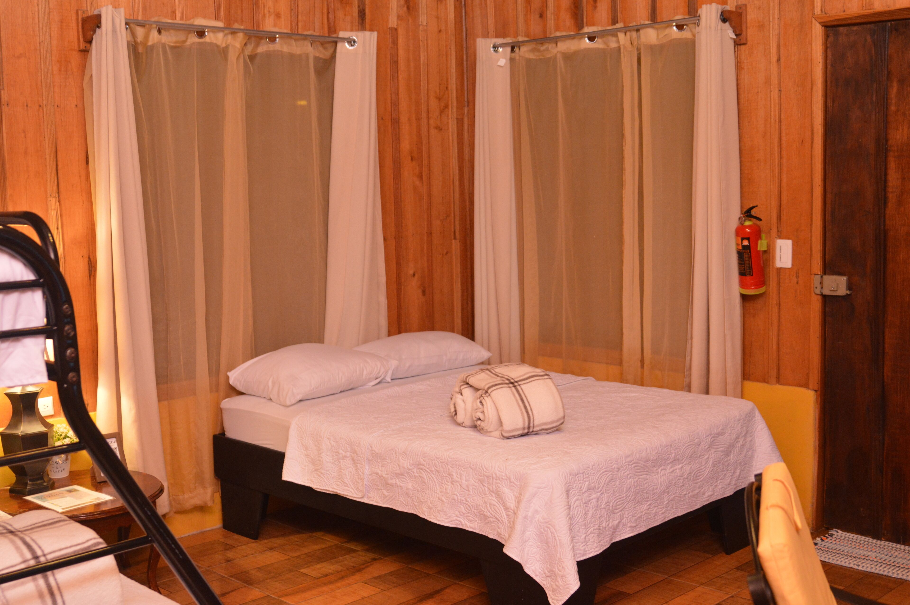 Una cama individual con una colcha y almohadas blancas, suelo de madera y una puerta de madera en una habitación con paredes de madera.