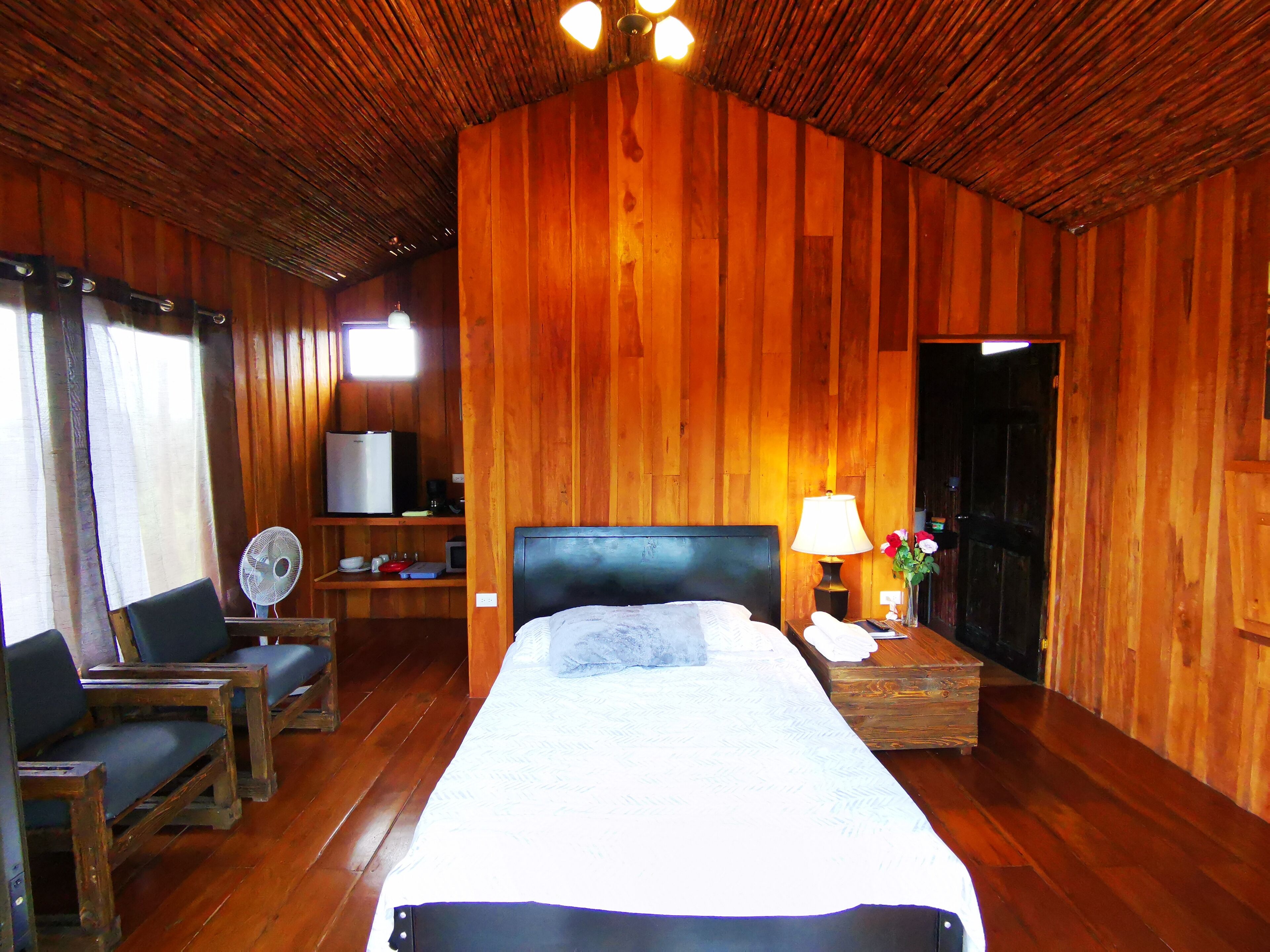 A wooden cabin room with a bed, a desk, a chair, a fan, and a television.