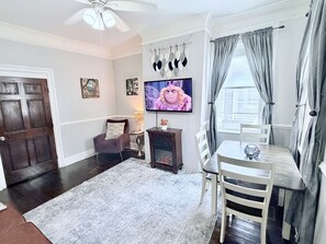 A living room with a fireplace, a television, a dining table, and a ceiling fan. - Historic Savannah Condo, Sleeps 6, Pool, Dogs Ok (Savannah)