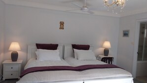 Un lit double avec du linge de lit blanc et des coussins décoratifs rouge foncé, deux tables de chevet avec des lampes, un ventilateur de plafond et un lustre.