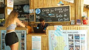 A check-in counter with a chalkboard displaying check-in and check-out times, a laptop, and a menu board with various activities. - Bodega Pool Party Koh Phangan (Ko Pha-ngan)