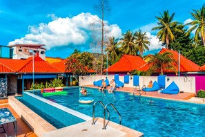 Una piscina con sillas reclinables azules, un puesto de socorrista y palmeras al fondo.