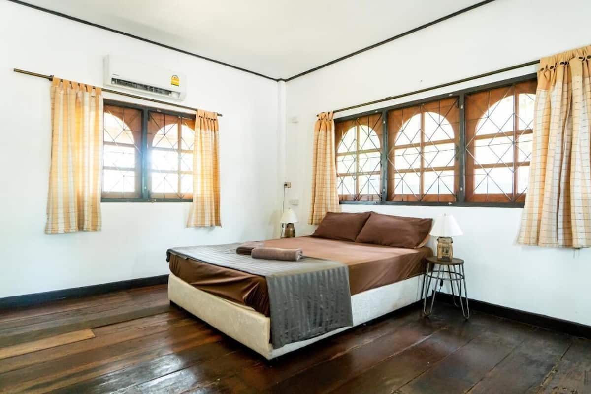 A bedroom with a bed, two windows with curtains, an air conditioning unit, and a small table with a lamp.