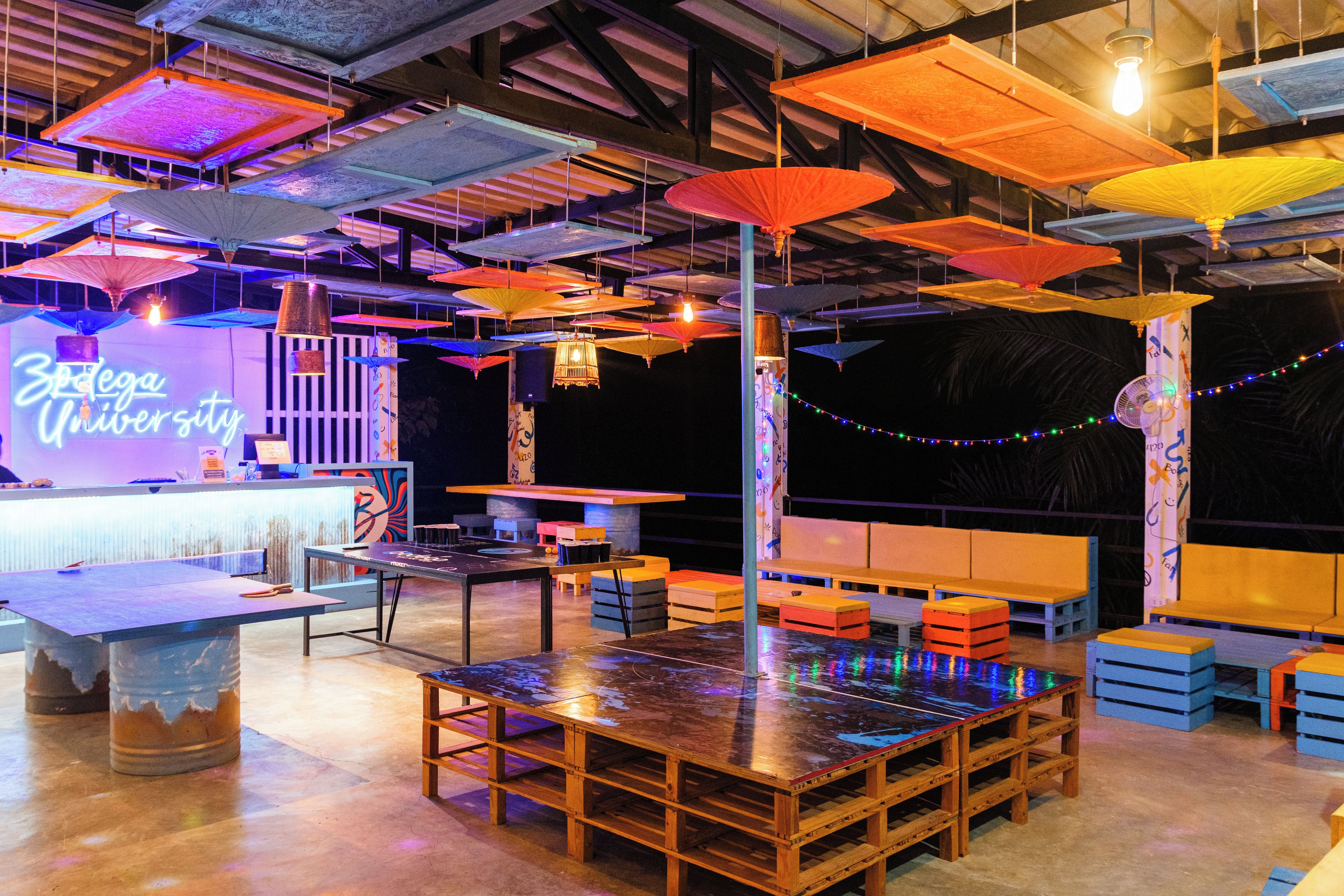 A spacious venue with tables and benches, a neon sign, and hanging umbrellas.