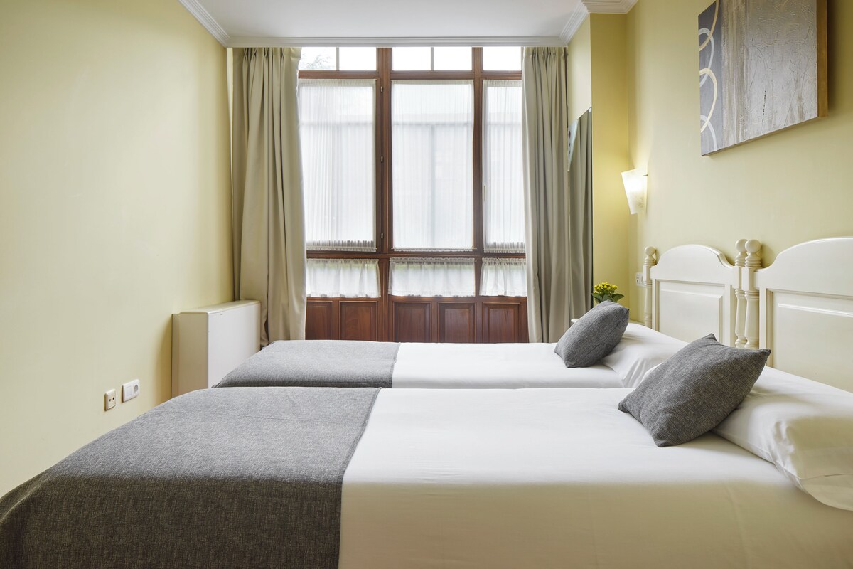 Habitación de hotel con dos camas, un ventanal con cortinas, una lámpara de pared y un cuadro.