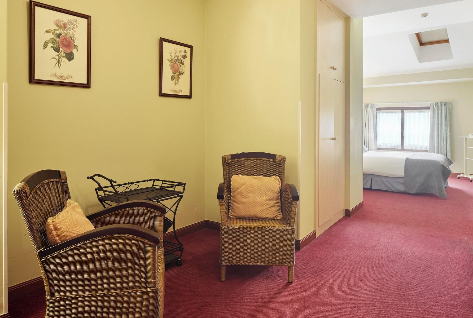Een hotelkamer met rieten stoelen, een bed en twee ingelijste bloemwerk afbeeldingen aan de muur.