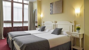 Habitación de hotel con dos camas, una mesita de noche con lámpara y una ventana con cortinas.
