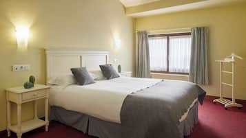 A hotel room with a bed, bedside tables, a nightstand, a window with curtains, and a coat rack.