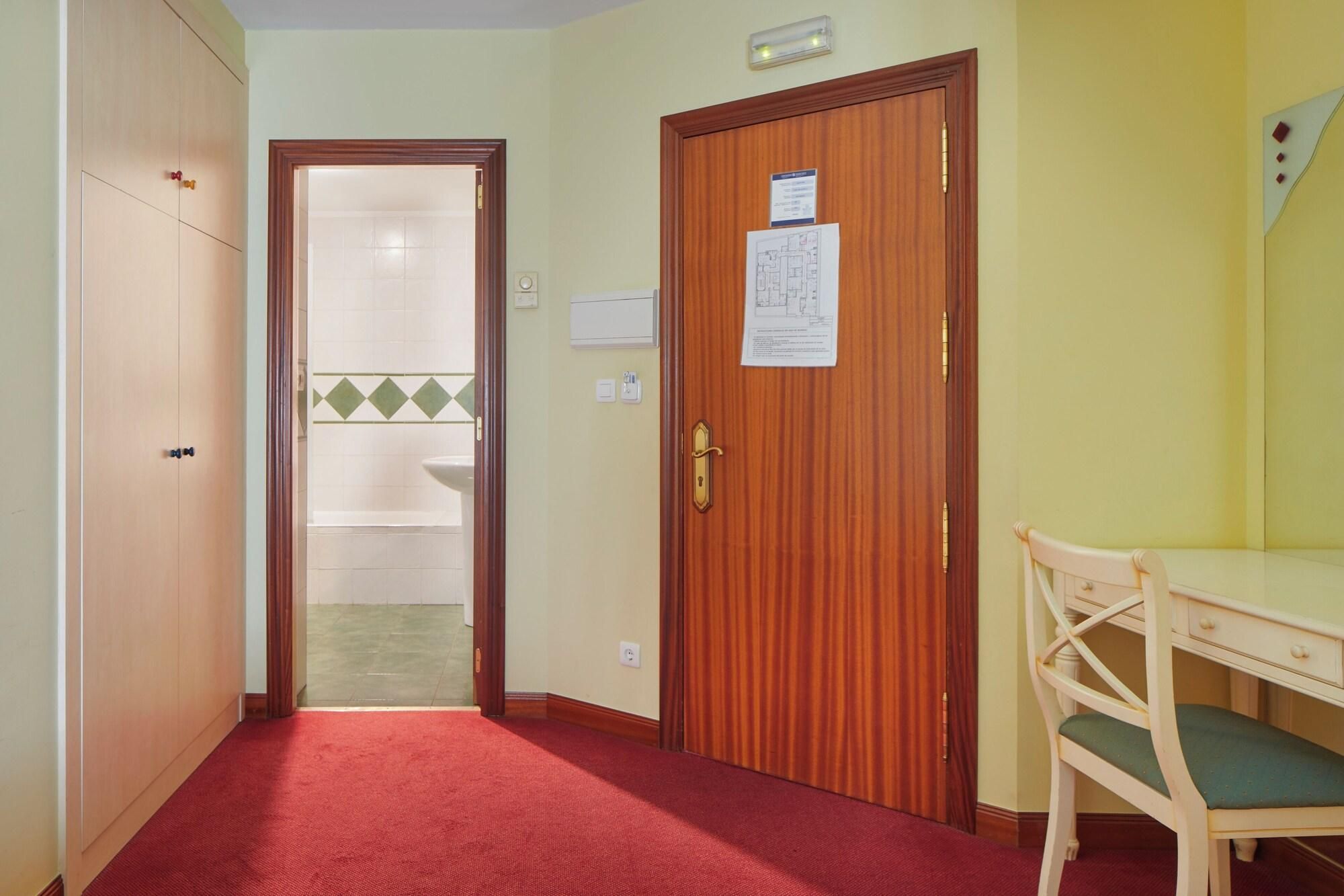 Habitación de hotel con puerta de madera, pasillo con alfombra roja, un escritorio de madera con una silla, y un baño visible a través de una puerta abierta.
