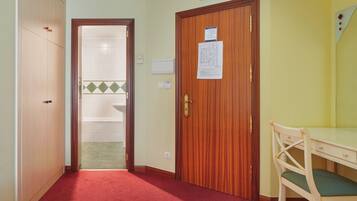Habitación de hotel con puerta de madera, pasillo con alfombra roja, un escritorio de madera con una silla, y un baño visible a través de una puerta abierta.