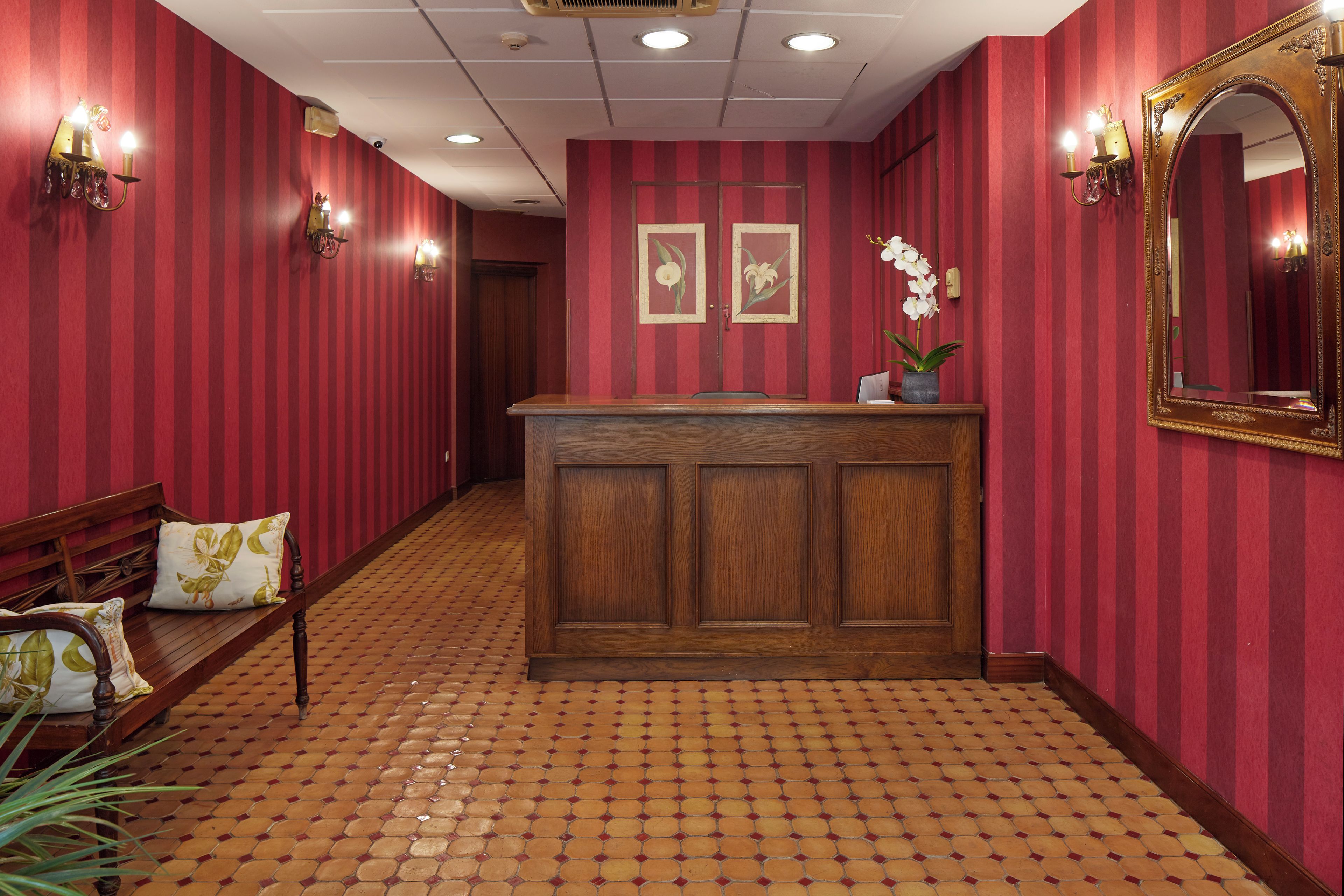 A hotel reception area with a wooden reception desk, red striped wallpaper, and a tiled floor.