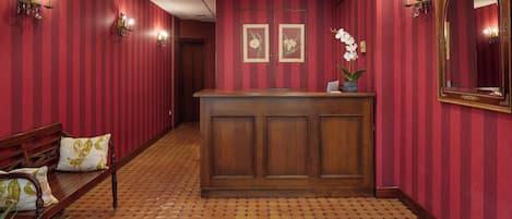 A hotel reception area with a wooden reception desk, red striped wallpaper, and a tiled floor.
