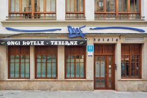 Fachada de hotel con amplios ventanales, un letrero destacado sobre la entrada y un toldo azul.