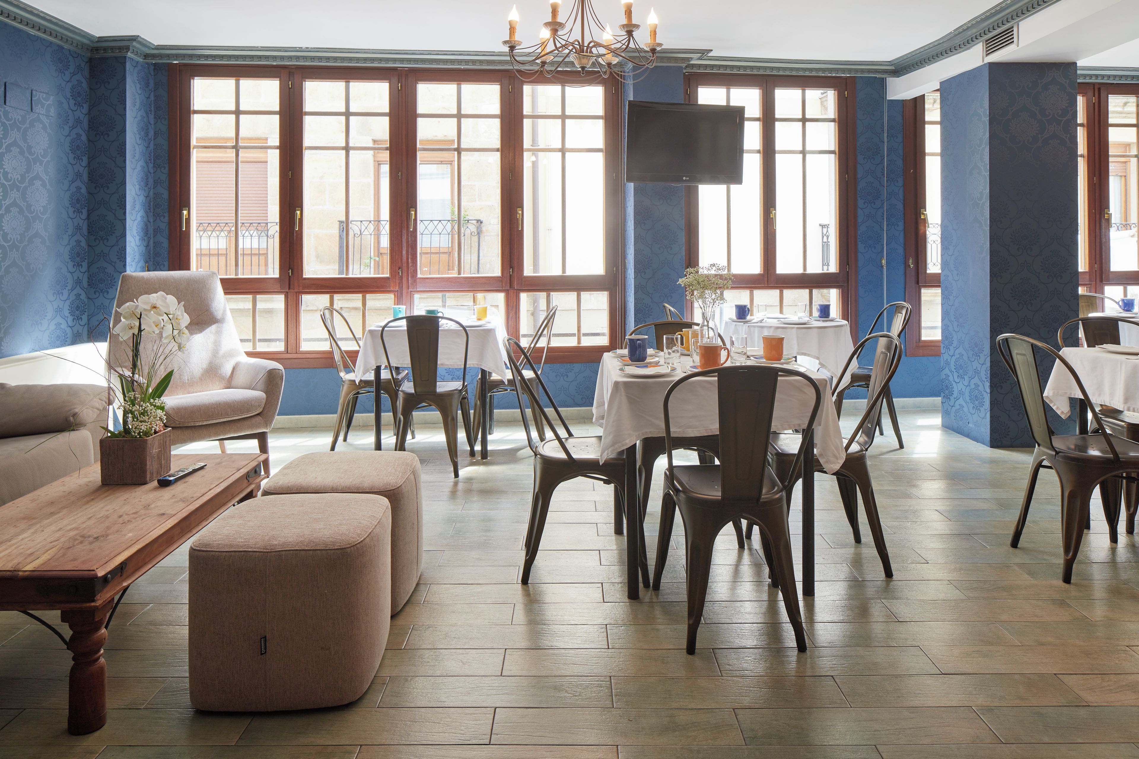 Ruime kamer met een groot raam, een bank, een salontafel en verschillende stoelen rondom een kleine tafel.