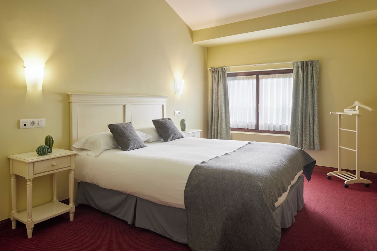 Une chambre d’hôtel comprenant un lit, des tables de chevet, une commode, une fenêtre avec des rideaux et un porte-manteau.