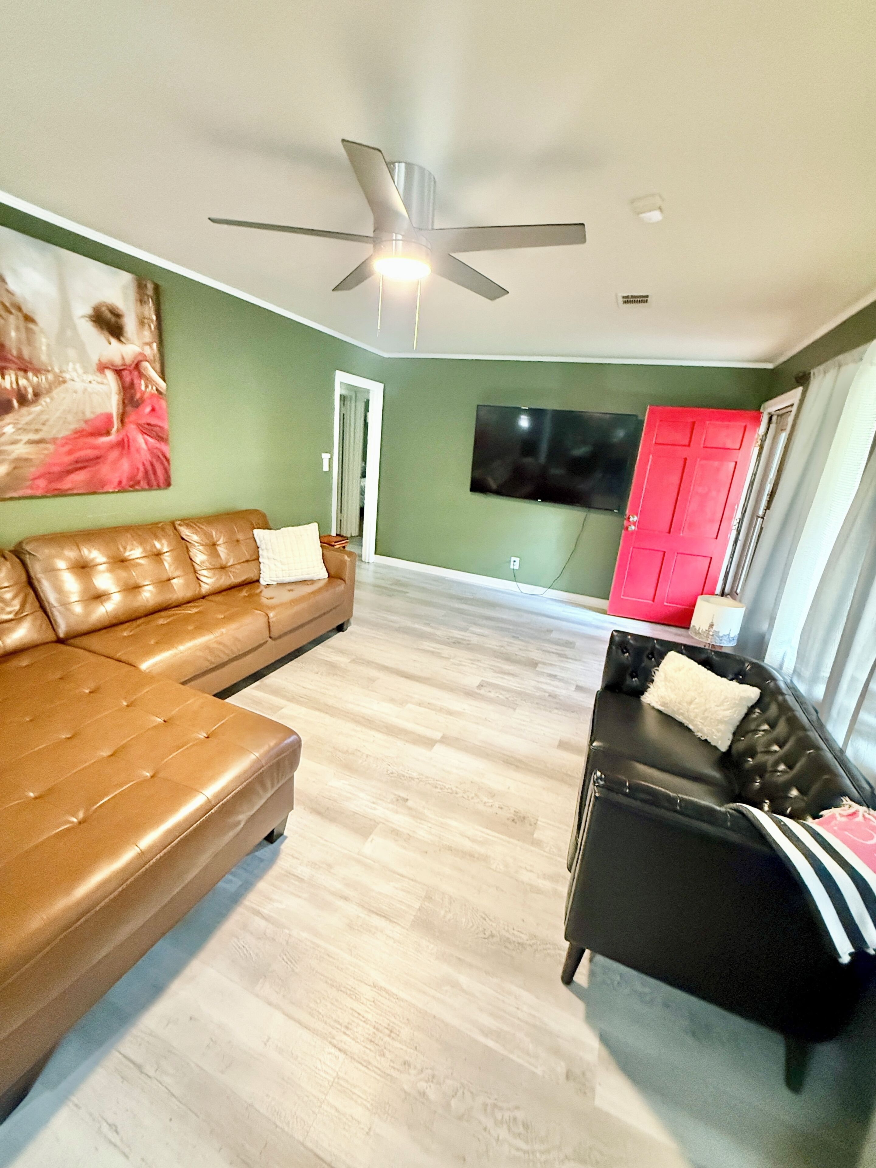 A living room with a brown leather sofa, a black leather sofa, a ceiling fan, a flat-screen TV, and a red door.