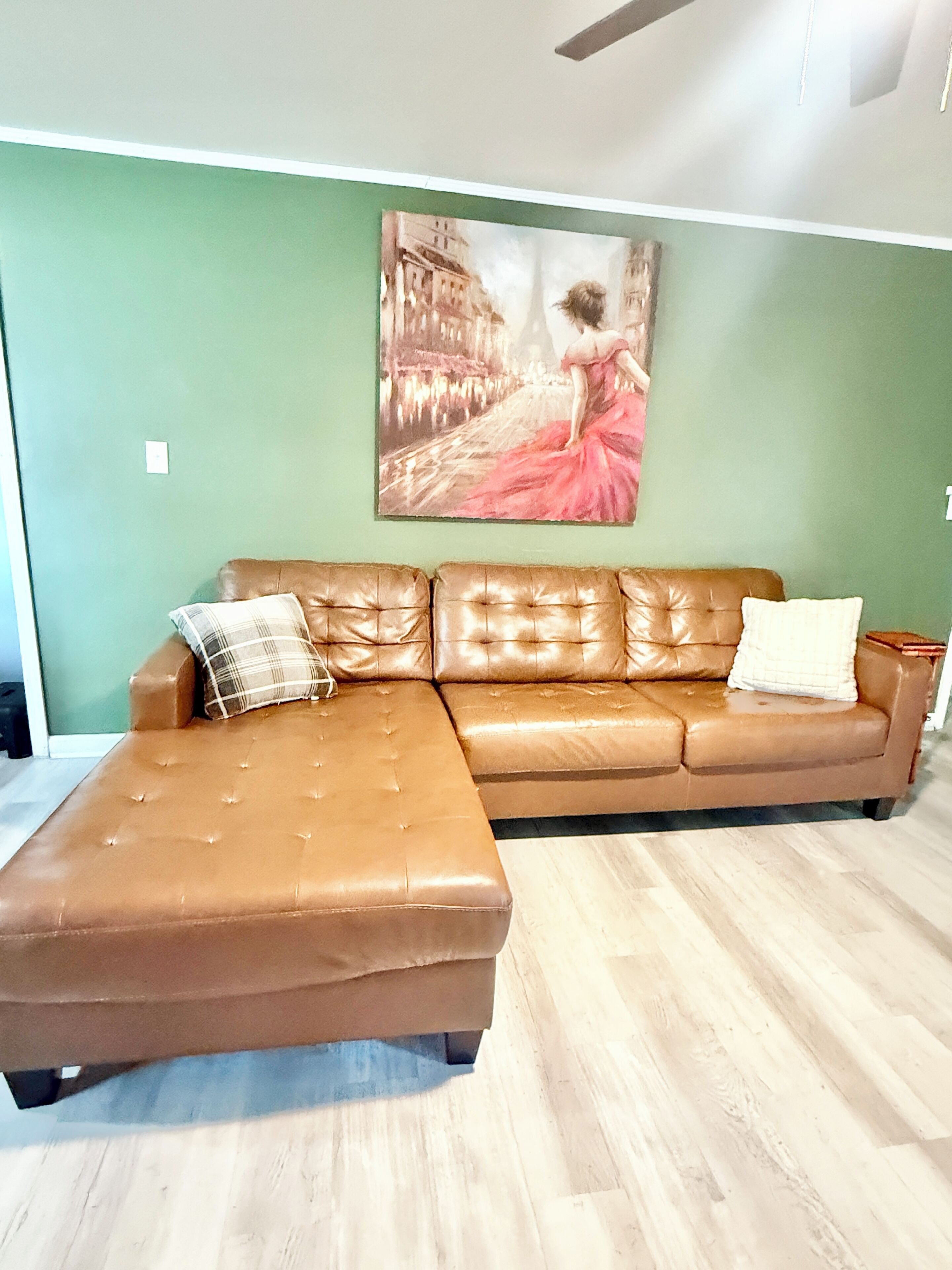 A brown leather sectional sofa with a chaise, a painting of a woman in a pink dress on the wall, and a light wooden floor.