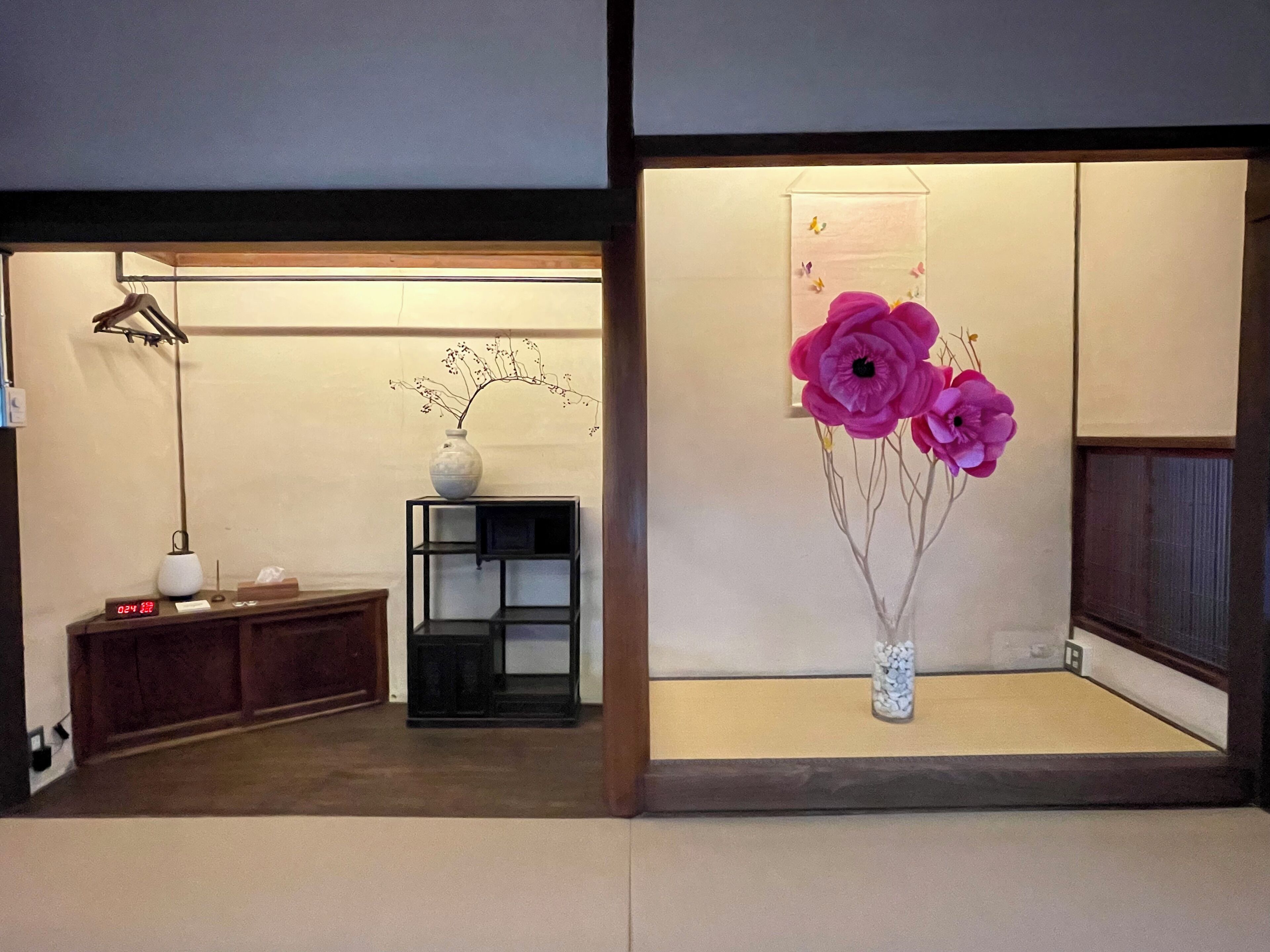 Una habitación con un armario de madera, un estante negro, un jarrón blanco con ramas y un arreglo de flores rosas.