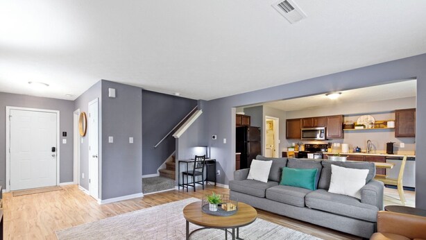 A modern living room with a grey sofa, a coffee table, and a staircase leading to a kitchen area. - Splendid 4 Bed/2.5 Bath- Near Speedway (Indianapolis)