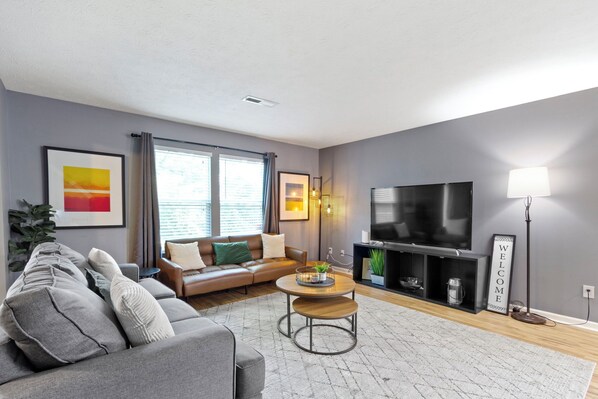 A modern living room with a grey sofa, a coffee table, a flat-screen TV, and a 'WELCOME' sign. - Splendid 4 Bed/2.5 Bath- Near Speedway (Indianapolis)