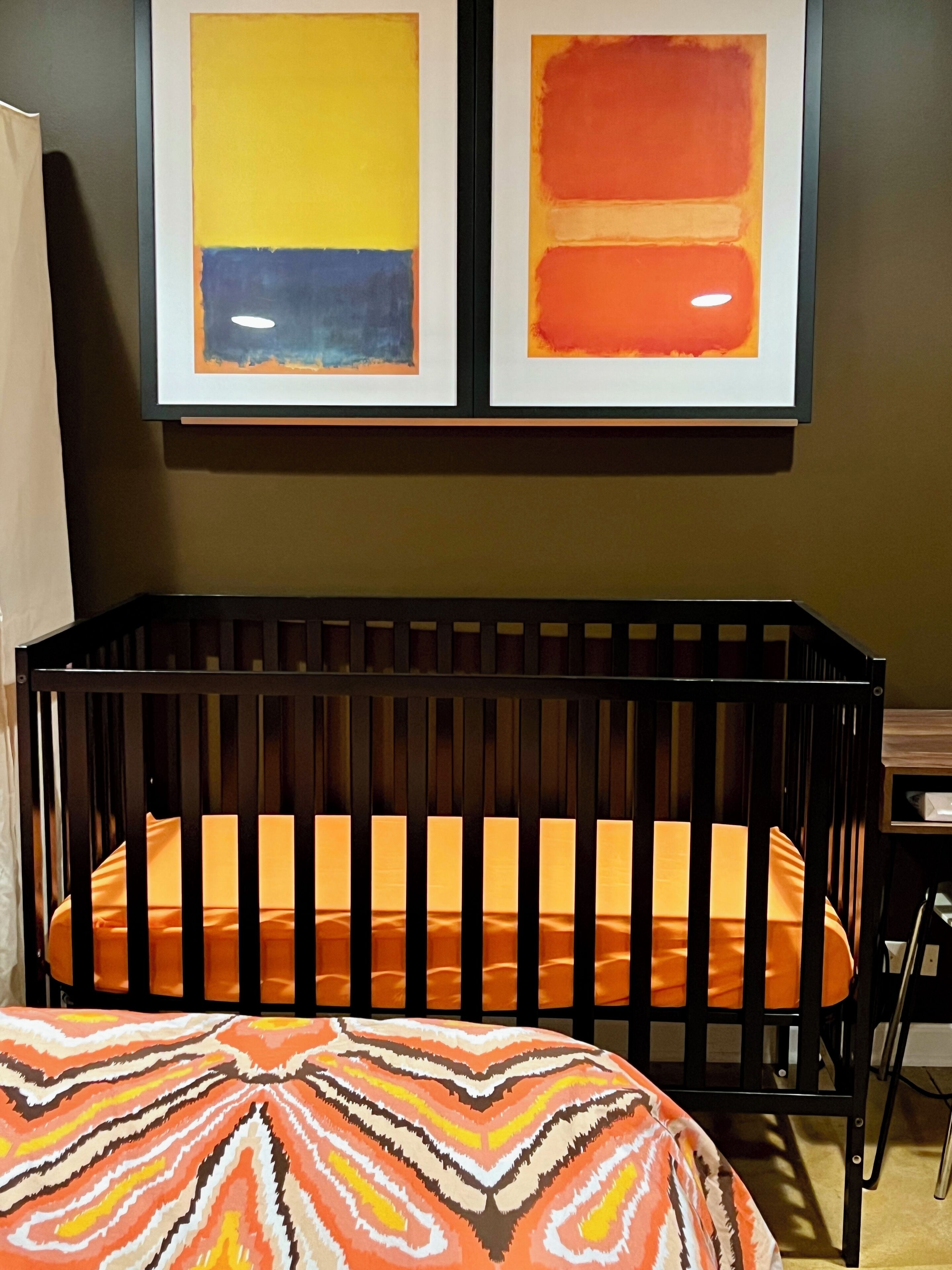 A crib with an orange mattress, a striped blanket, and two framed abstract paintings on the wall above.