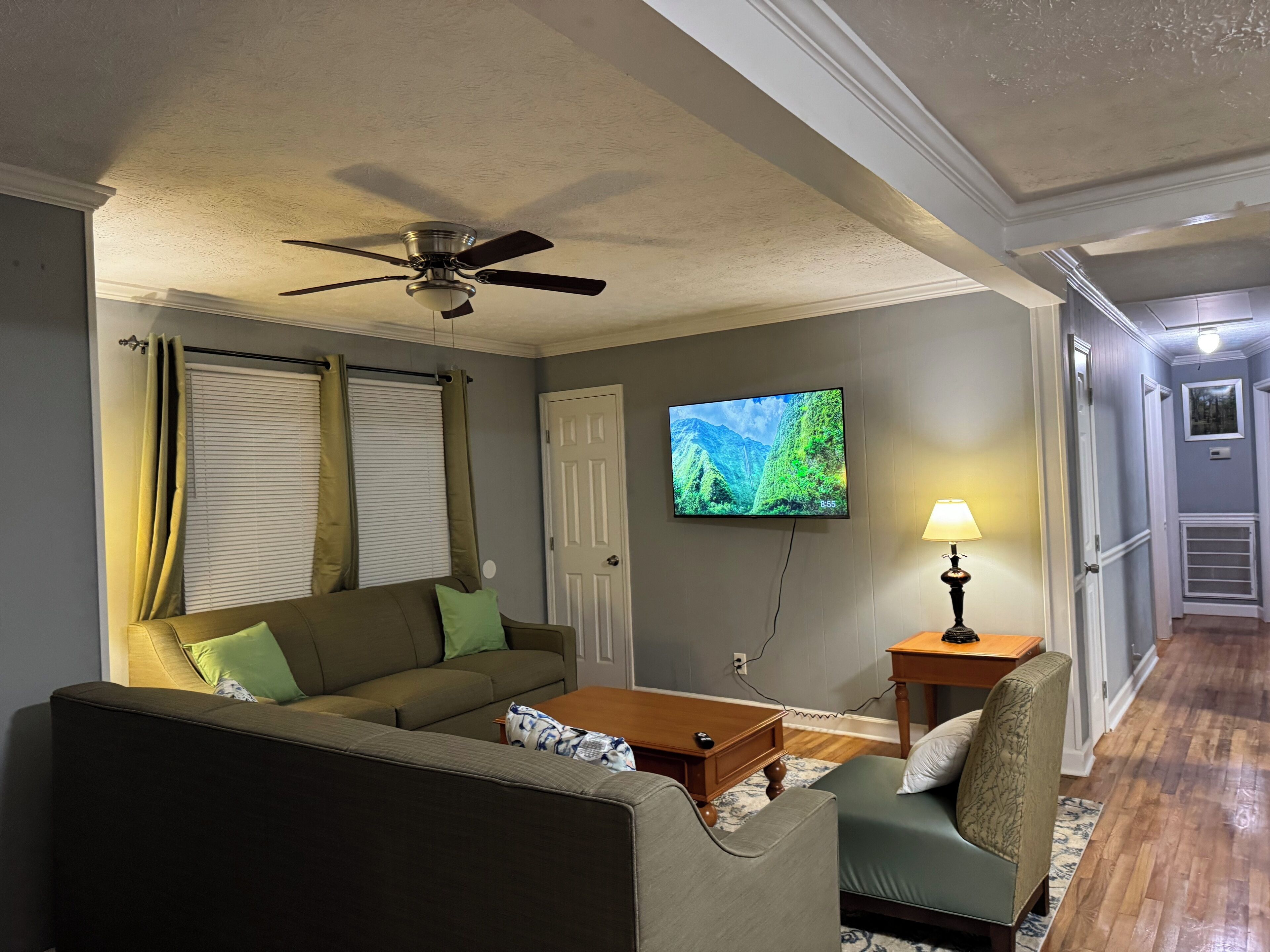 A living room with a sofa, a coffee table, a television mounted on the wall, a ceiling fan, and a lamp.