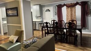 A dining area with a wooden table and chairs, a kitchen in the background, and a living room with a sofa and cushions. - Mountain View Home (Cave Spring)