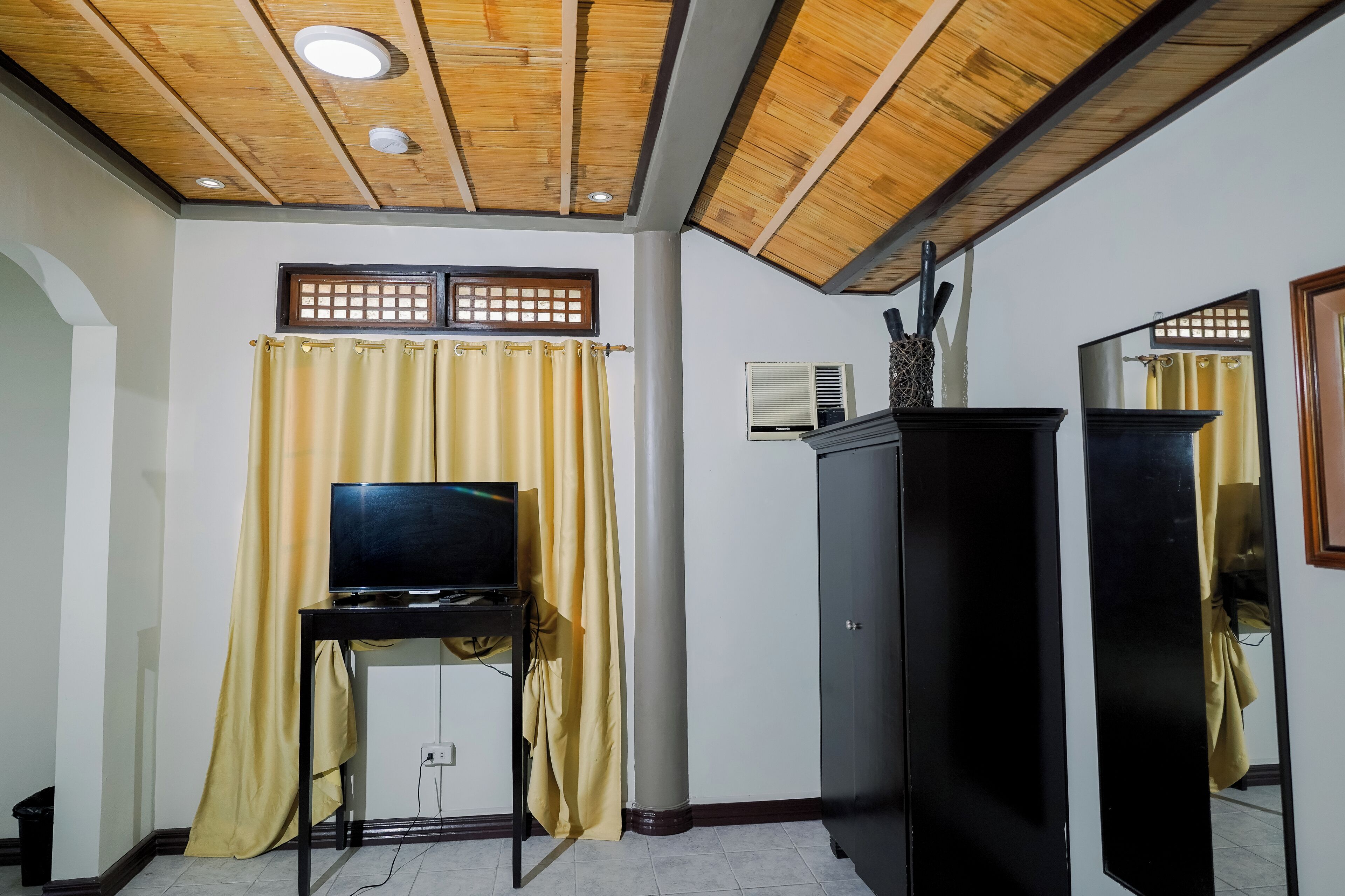 A room with a wooden ceiling, a television on a stand, a cabinet, and a window with curtains.
