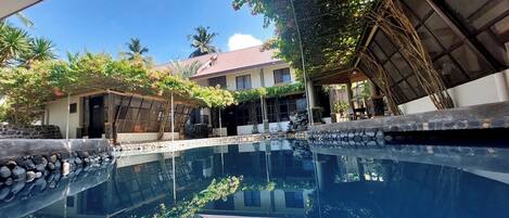 A swimming pool with a stone edge, surrounded by a covered patio area with a pergola and plants.