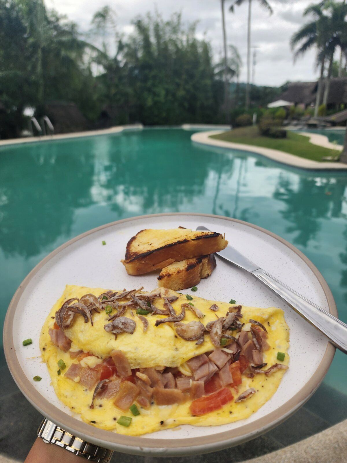 A breakfast plate with eggs, mushrooms, and ham, and toast, with a pool and greenery in the background.
