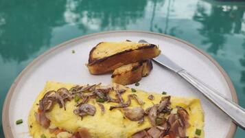 A breakfast plate with eggs, mushrooms, and ham, and toast, with a pool and greenery in the background.