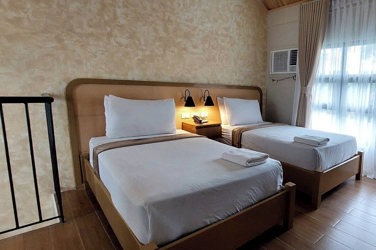 A double room with two beds, a bedside table with a lamp, an air conditioning unit, and a window with curtains.