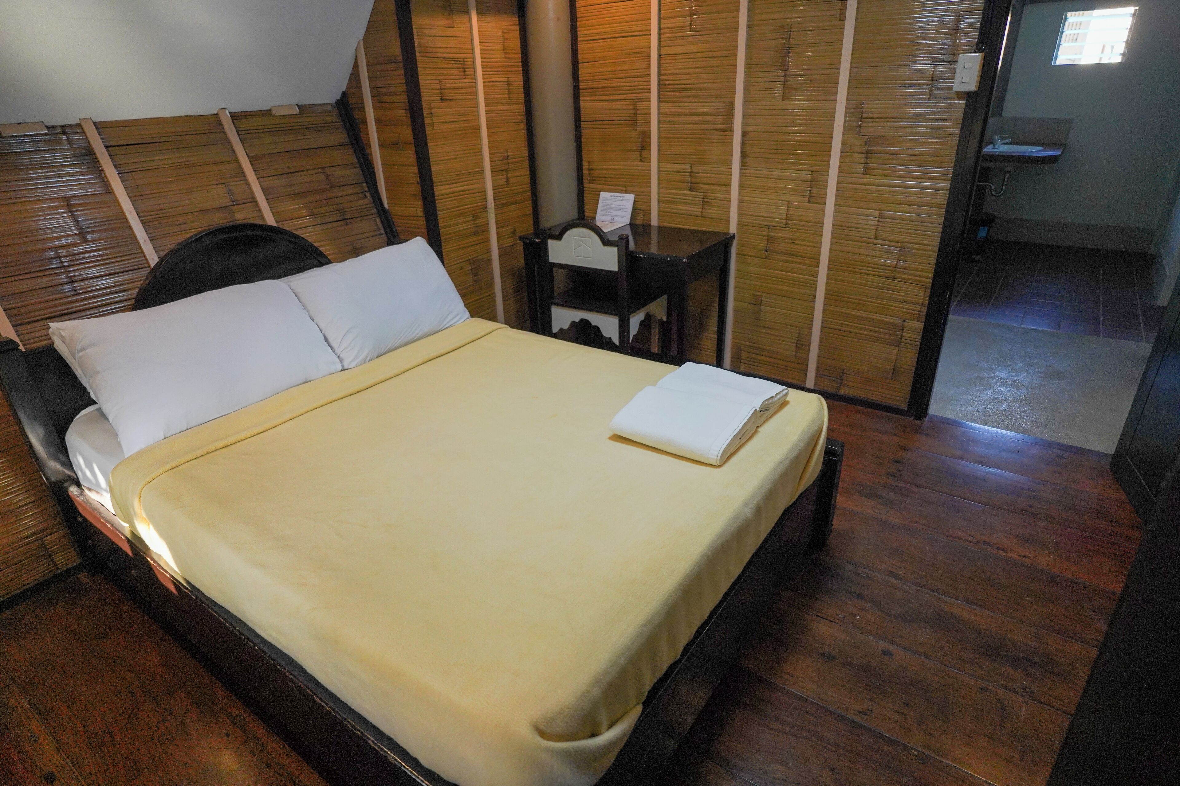 Una cama bien hecha con una almohada blanca y una colcha color beige, una mesita de noche de madera con una lámpara y suelo de madera.