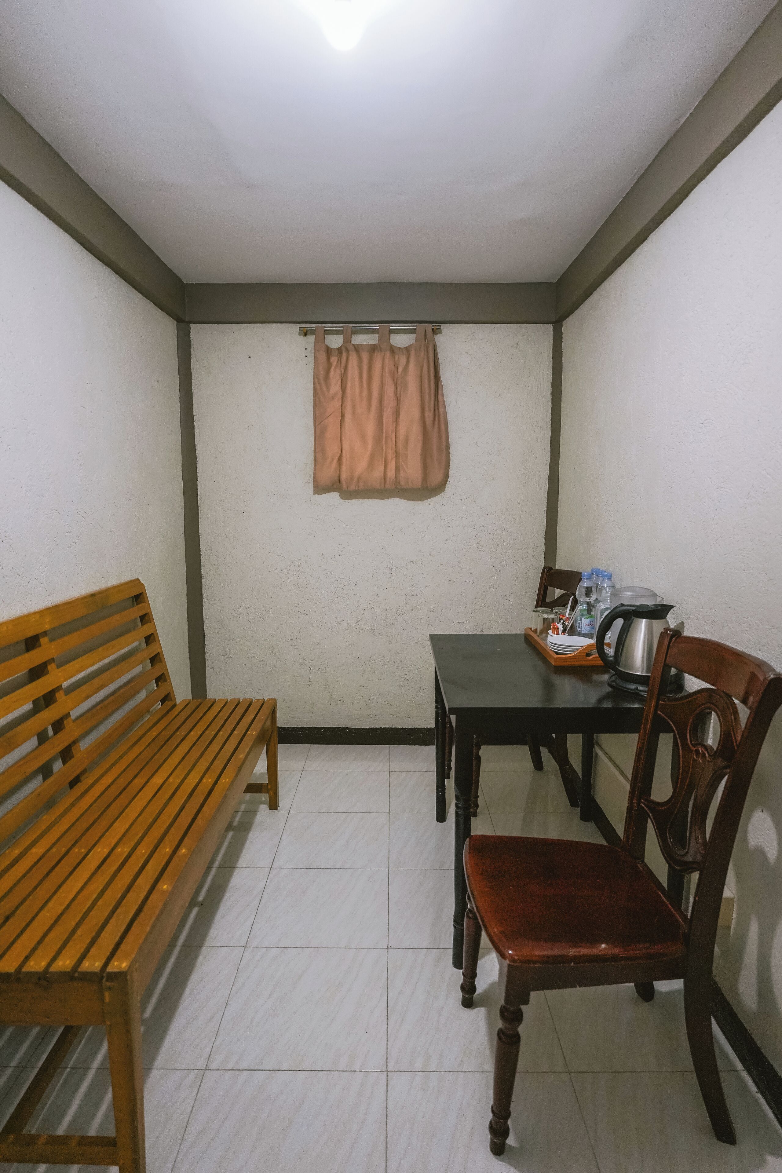 Una habitación pequeña y limpia con un banco de madera, una mesa con sillas y una ventana con cortinas.