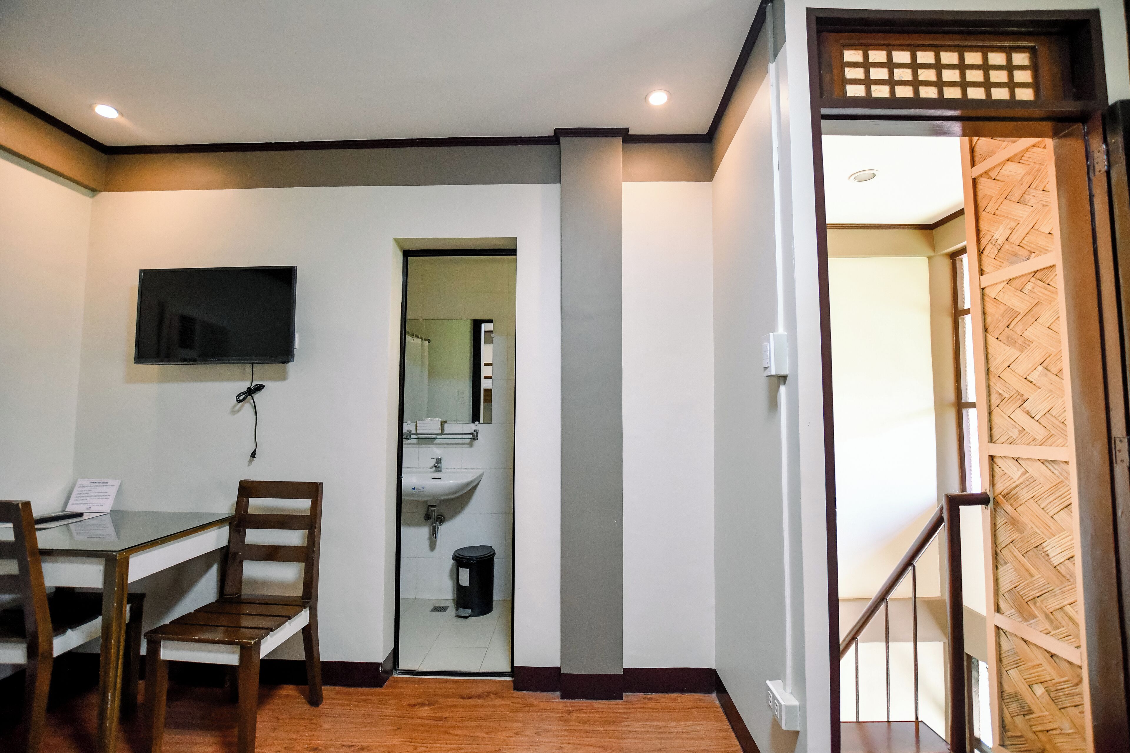 A room with a dining table, chairs, a TV mounted on the wall, a bathroom visible through an open door, and a wooden staircase.