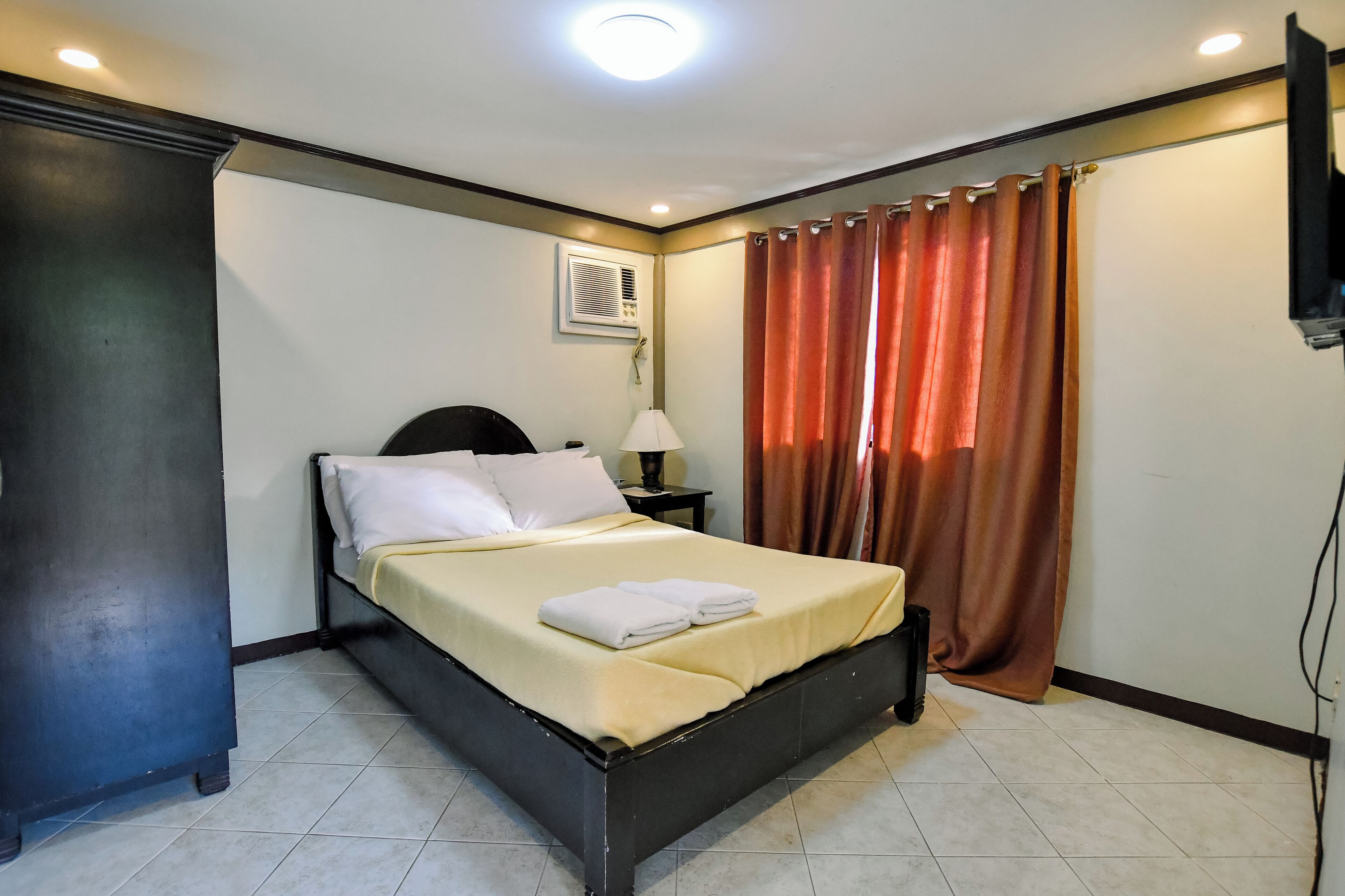A neatly made bed with pillows and a folded towel, a bedside lamp, and a wall-mounted air conditioner.