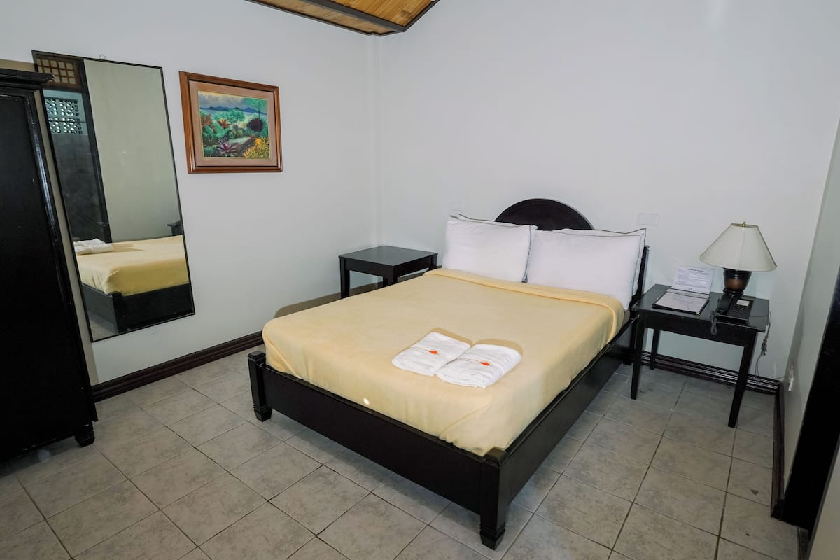 A neatly made bed with a beige bedspread and two towels, a small bedside table with a lamp, a nightstand, a mirror, and a framed painting on the wall.
