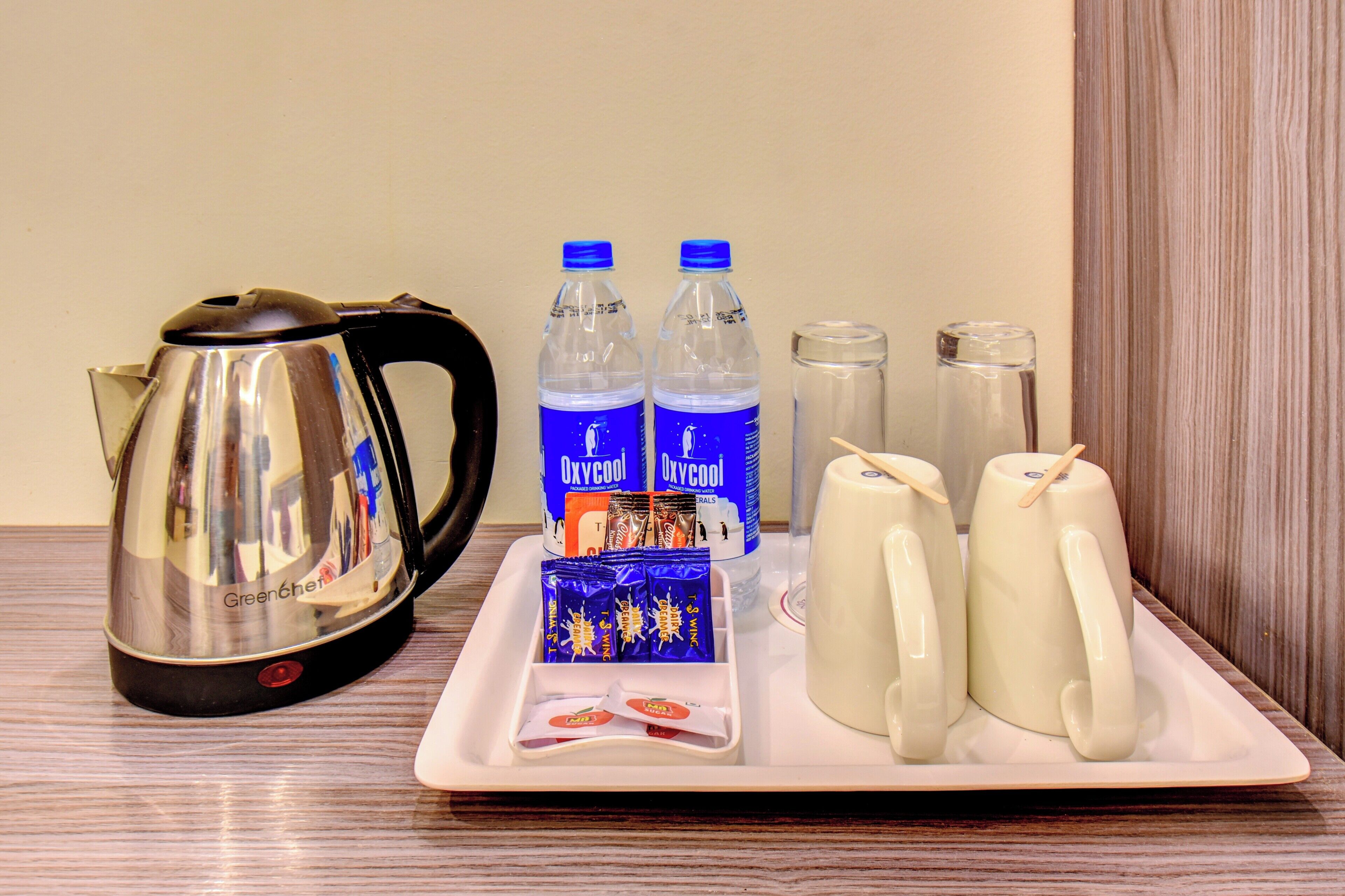 A tray with a kettle, two water bottles, a box of snacks, and two mugs on a wooden table.