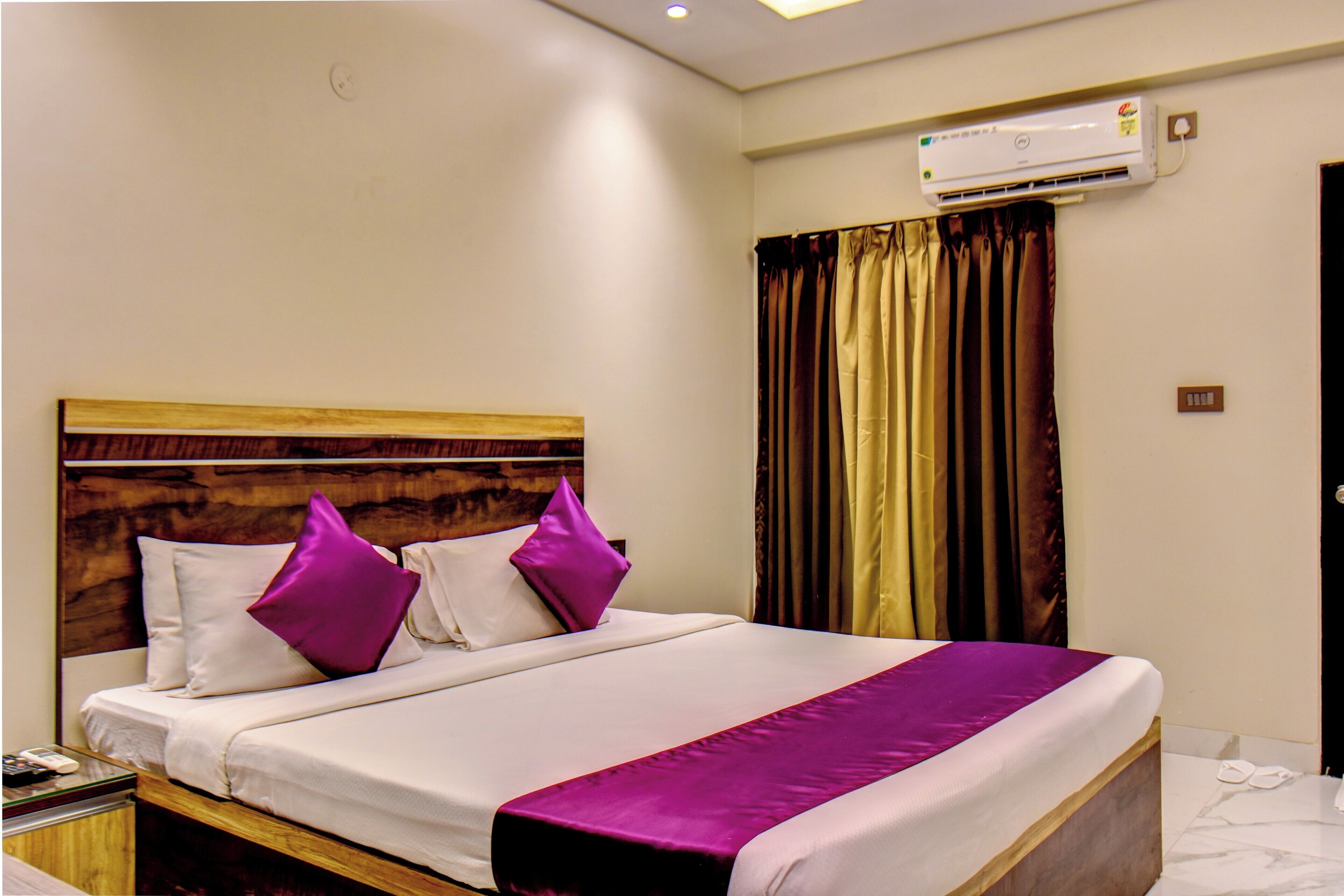 Une chambre d’hôtel avec une tête de lit en bois, un lit avec une literie blanche et des touches de couleur violette, un climatiseur fixé au mur et un rideau.