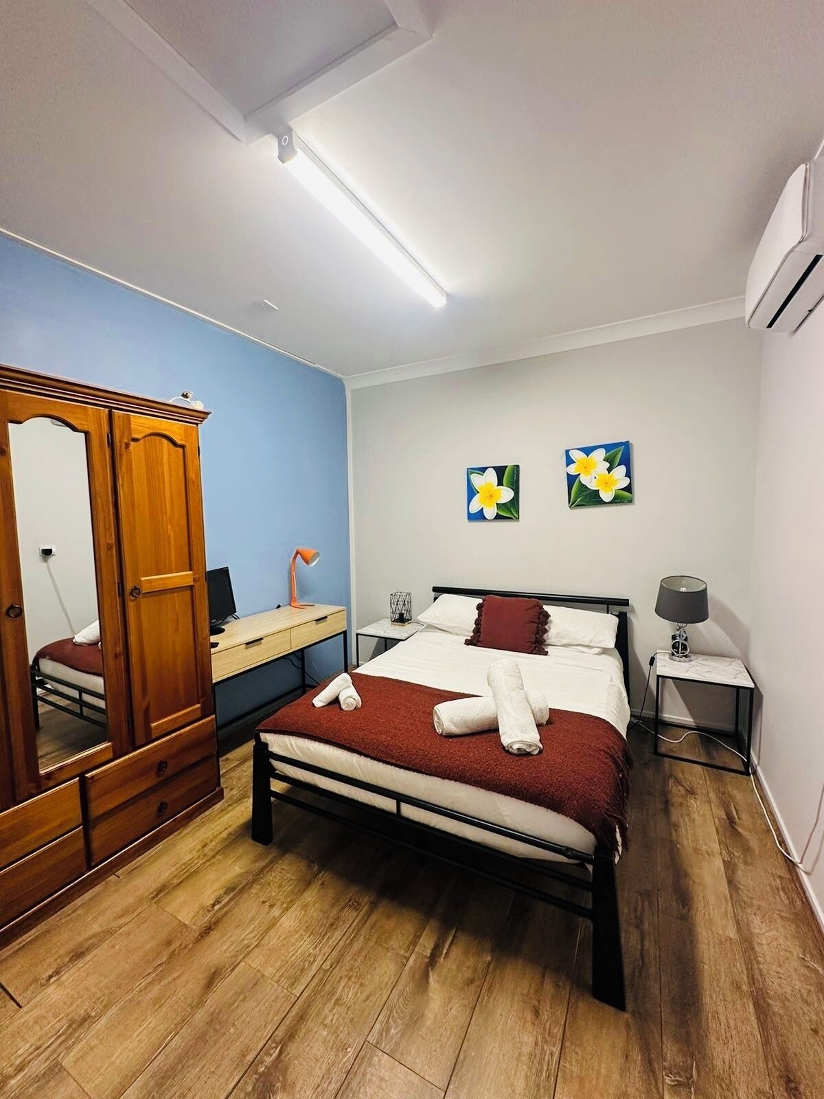 Un dormitorio con una cama, un armario de madera, un escritorio, una silla, un espejo y dos cuadros de flores colgados en la pared.