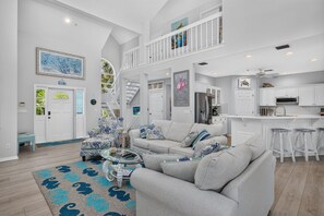 Amplia sala de estar con techos altos, una escalera que conduce a un nivel superior y una cocina con muebles blancos.