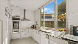 A modern kitchen with white cabinetry, stainless steel appliances, a large window, and a view of a house. - Lake Taupo Gem (Taupo)