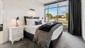 A bedroom with a large bed, a nightstand, a lamp, and a view of the outside. - Lake Taupo Gem (Taupo)
