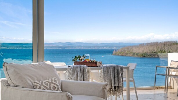 A balcony with a view of a lake, furnished with a sofa, chairs, and a table with a fruit basket.