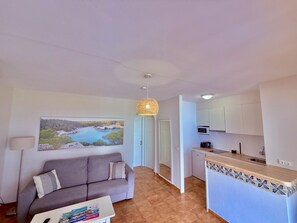 A living room with a sofa, a coffee table, a painting of a coastal landscape, and a kitchen with a microwave and cabinets. - Enjoy Cala Porter - Apto en Complejo Vacacional (Cala'N Porter)